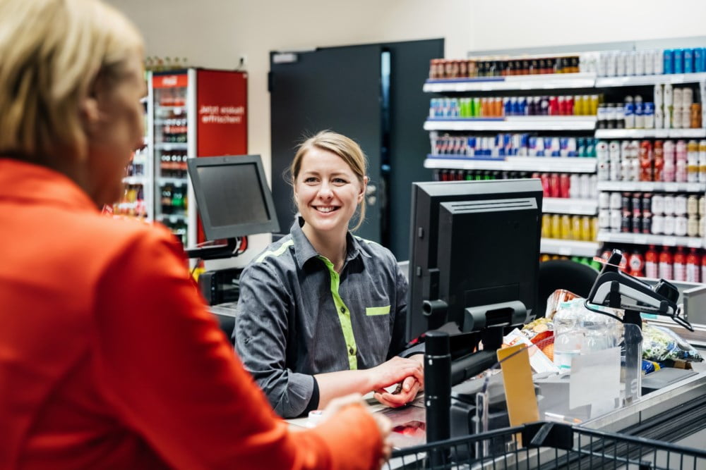Smilende butikkmedarbeider ved kassen i en dagligvarebutikk, mens en kunde håndterer kontanter
