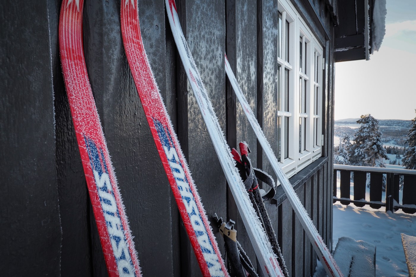 Frostdekkede røde og hvite langrennsski som står inntil en svart hyttevegg
